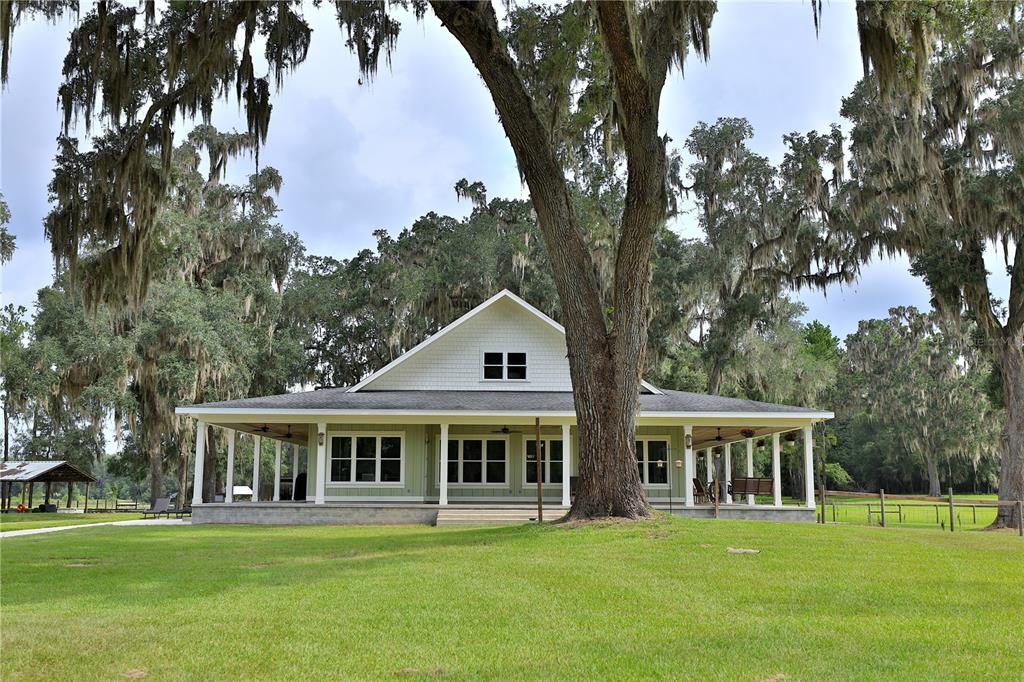 A light green house with a wrap-around porch sits on a grassy lawn under trees — 8410 NE 155TH AVENUE, WILLISTON, FL