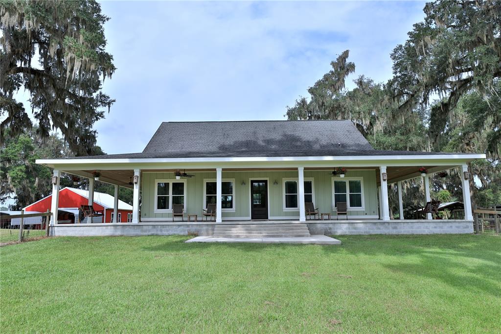 A light green house with a wrap-around porch stands on a grassy lawn under trees — 8410 NE 155TH AVENUE, WILLISTON, FL