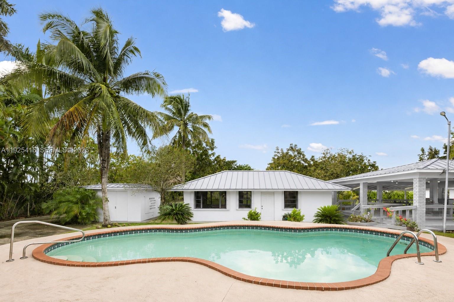 A kidney-shaped swimming pool sits in front of a white house with a metal roof — 20450 SW 198th Ave, Miami FL 33187, MIAMI, FL
