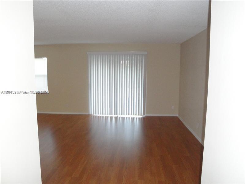 An empty room features light-colored walls, wood-look flooring, and vertical blinds over a sliding door — 18111 NW 68th Ave # I208, Hialeah FL 33015, HIALEAH, FL