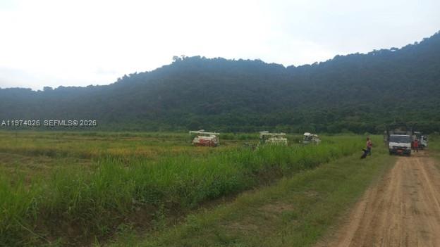 A dirt road runs alongside a field with farm machinery and a forested mountain backdrop — KM 40 VIA NARANJAL-GUAYAS, EC, EC