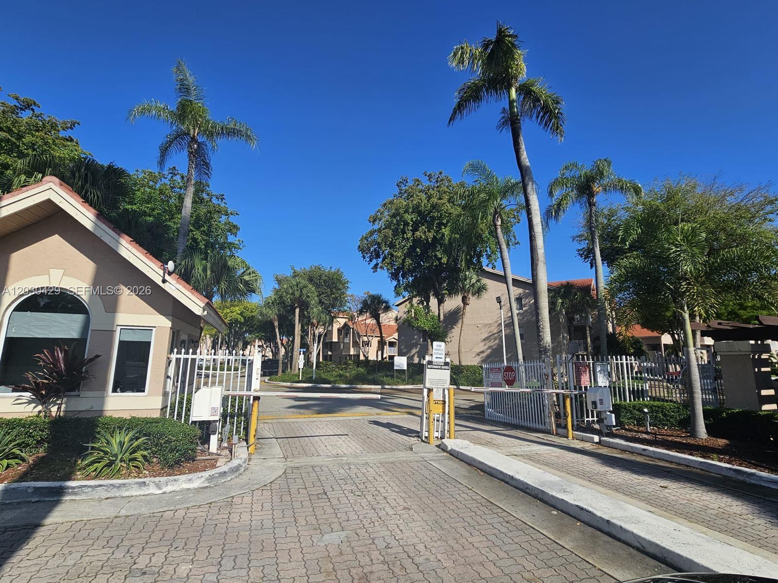 A gated entrance with a guardhouse, palm trees, and a paved driveway under a blue sky — 15771 SW 106th Ter # 204, Miami FL 33196, MIAMI, FL