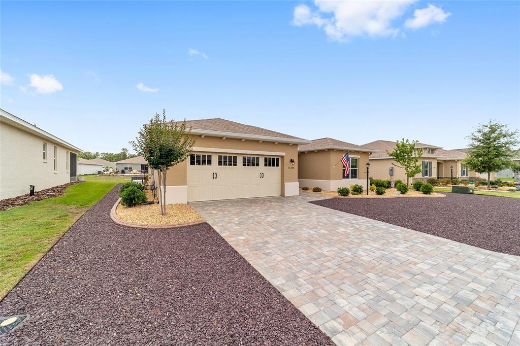 A tan single-story house features a two-car garage, a paver driveway, and landscaped rock beds — 10344 SW 98TH PLACE ROAD, OCALA, FL