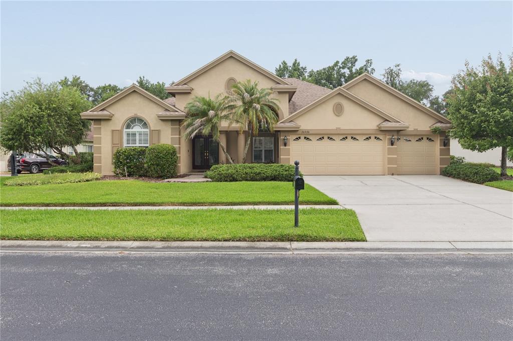 A tan single-story house with a three-car garage, palm trees, and a manicured lawn — 26920 WINGED ELM DRIVE, WESLEY CHAPEL, FL