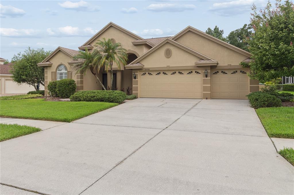A tan, single-story suburban house with a three-car garage and a concrete driveway — 26920 WINGED ELM DRIVE, WESLEY CHAPEL, FL