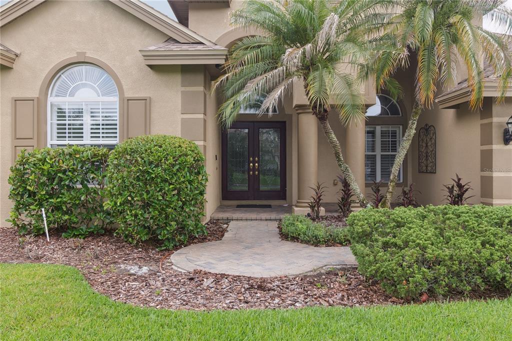 A beige house exterior features a paved walkway leading to a dark double front door — 26920 WINGED ELM DRIVE, WESLEY CHAPEL, FL