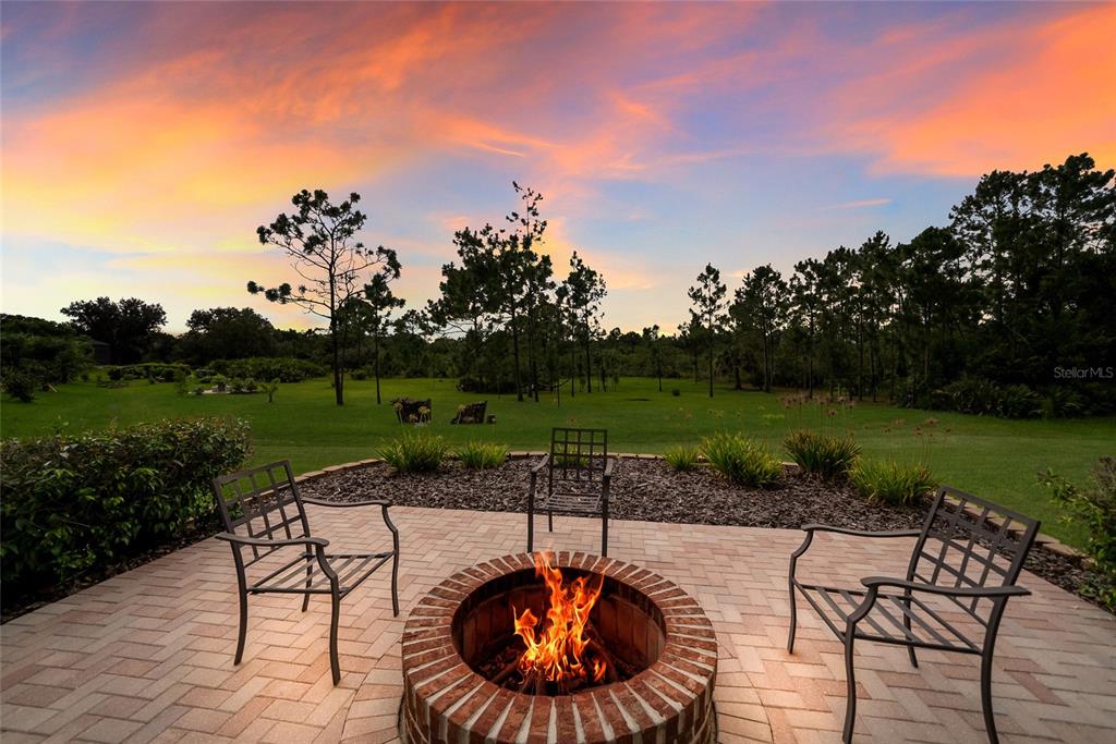 A brick fire pit sits on a patio with three chairs overlooking a grassy backyard — 23327 RED ROBIN PLACE, BRADENTON, FL
