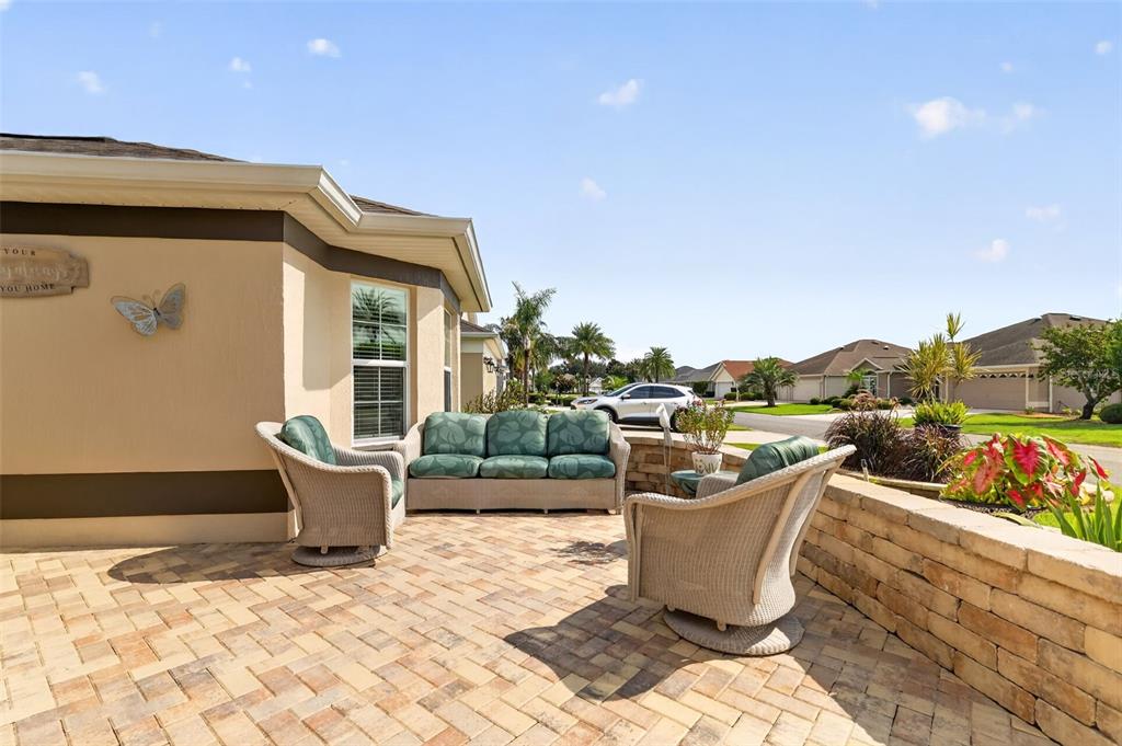 A paved patio features outdoor wicker furniture set against a house exterior with a stone wall — 2947 RAIN LILY LOOP, THE VILLAGES, FL