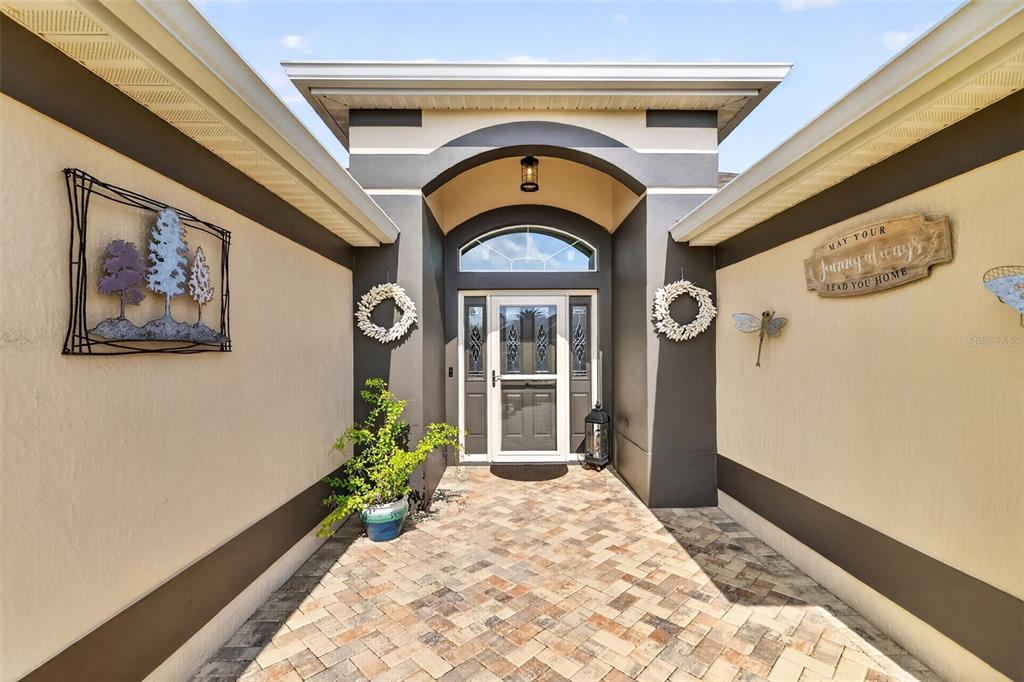A front entryway features a brick paver walkway, a dark arched doorway, and decorative wall art — 2947 RAIN LILY LOOP, THE VILLAGES, FL