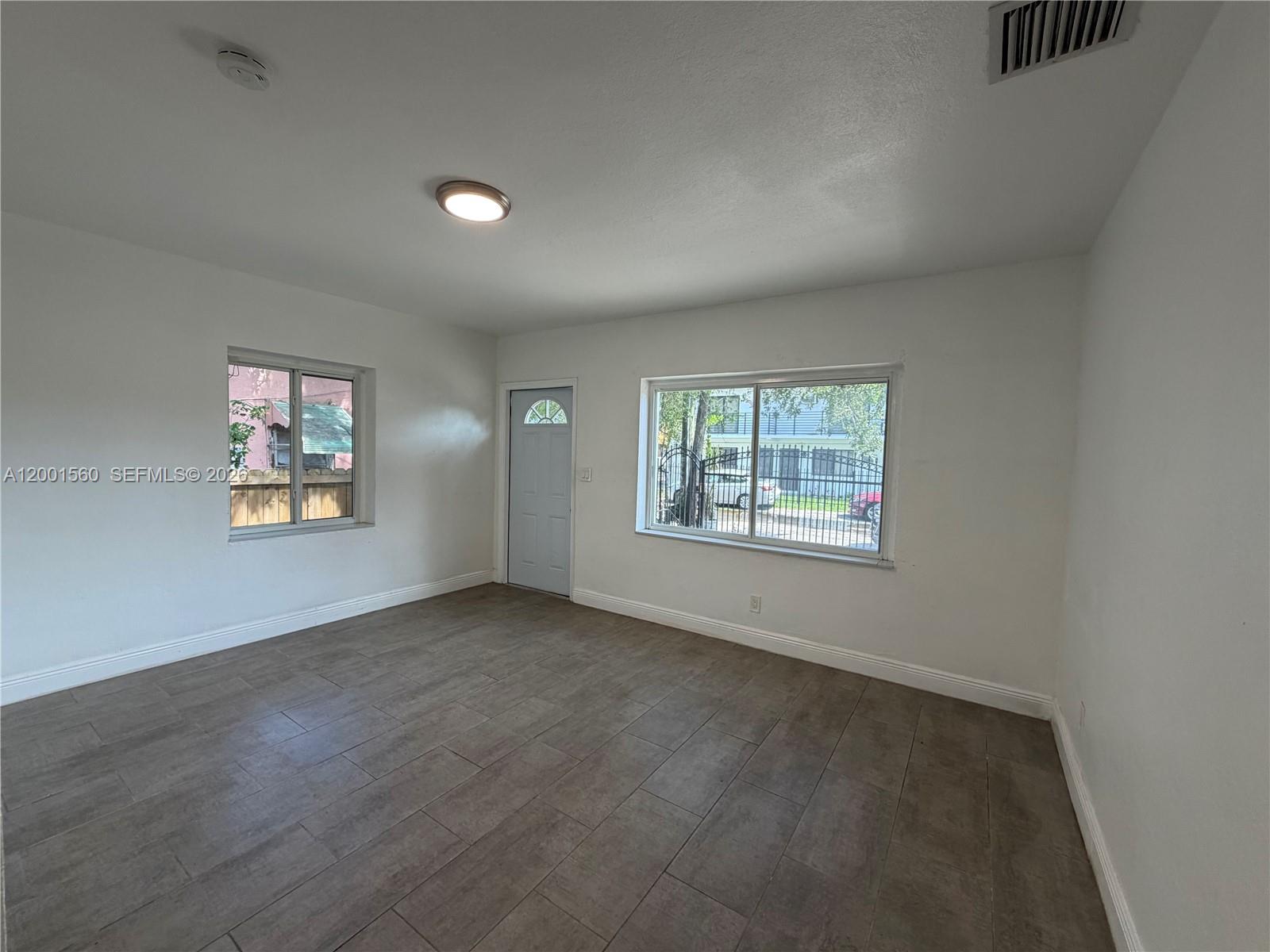 An empty room features white walls, gray tile flooring, two windows, and a single door — 7712 NE 1st Ct, Miami FL 33138, MIAMI, FL