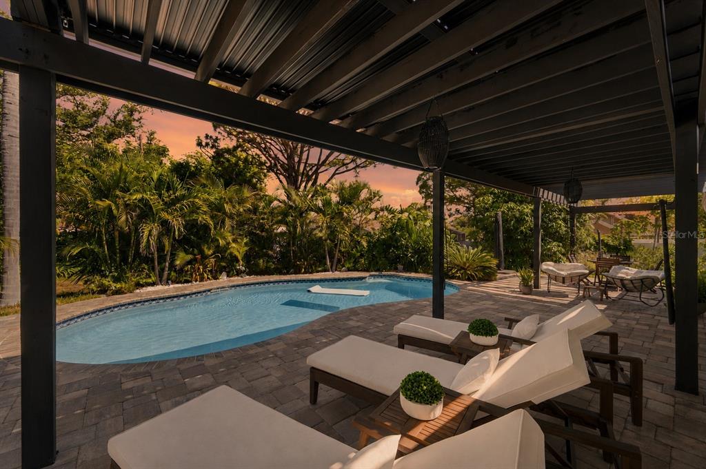 A covered patio with lounge chairs overlooks a swimming pool surrounded by lush tropical landscaping — 14297 GENEVA DRIVE, LARGO, FL