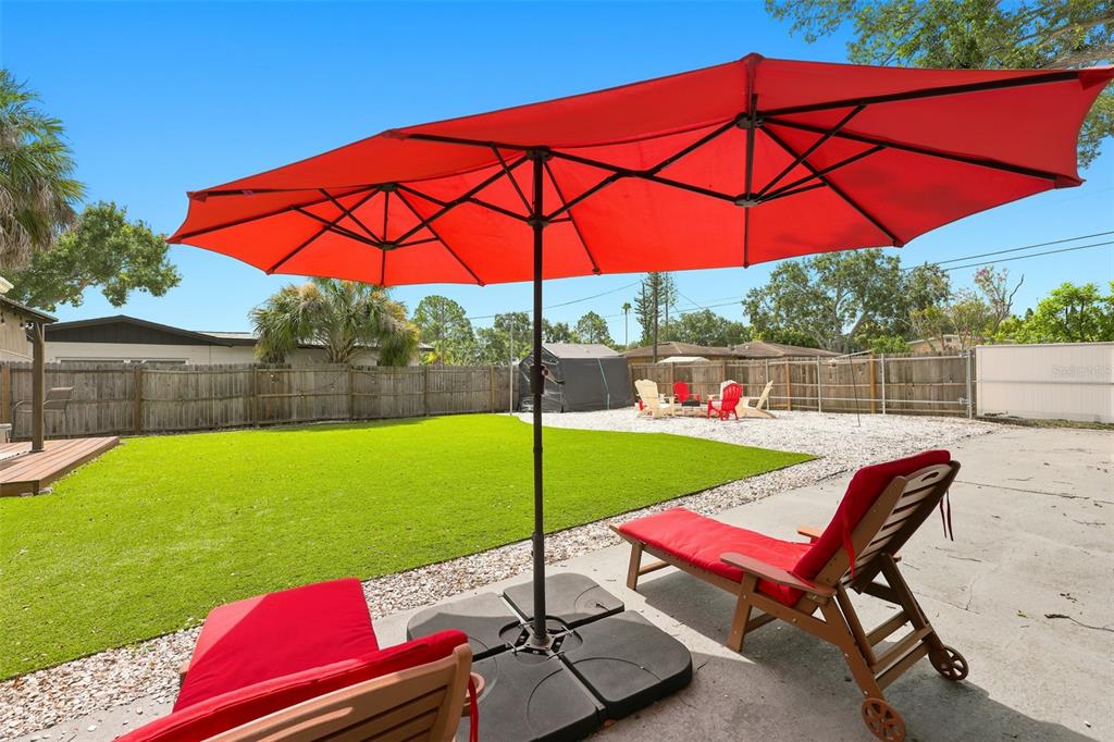 A backyard features a red double umbrella over lounge chairs, artificial turf, and a gravel area — 716 75TH AVENUE N, ST PETERSBURG, FL
