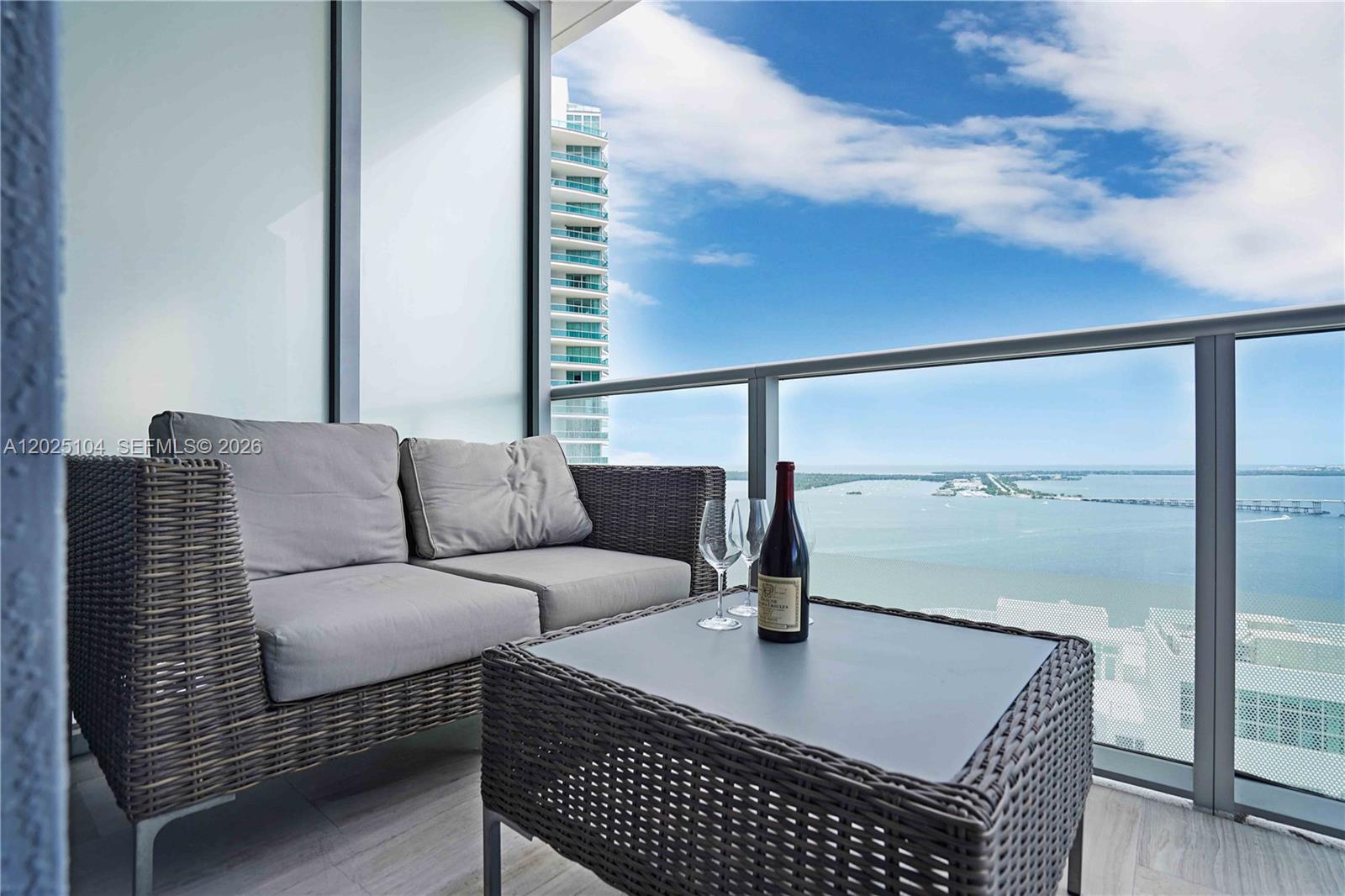 A balcony features a wicker sofa, a matching table with wine, and a water view — 1300 Brickell Bay Dr # 3402, Miami FL 33131, MIAMI, FL
