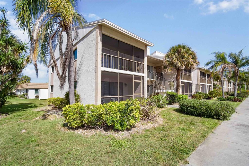 A two-story apartment building features screened-in balconies, light-colored stucco walls, and surrounding palm trees — 5476 SWIFT ROAD UNIT 19, SARASOTA, FL
