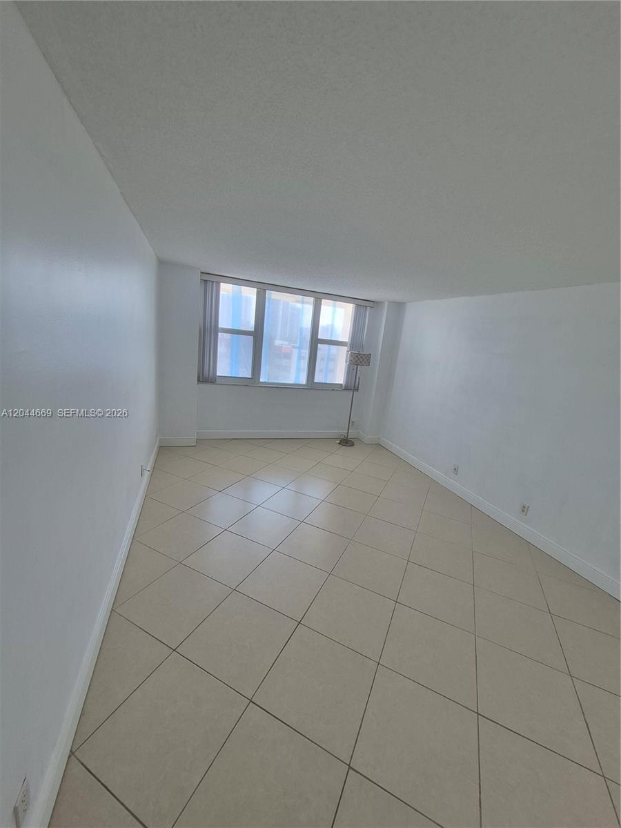 An empty room features beige tile flooring, white walls, a hanging chandelier, and a window — 3180 S Ocean Dr # 621, Hallandale Beach FL 33009, HALLANDALE BEACH, FL