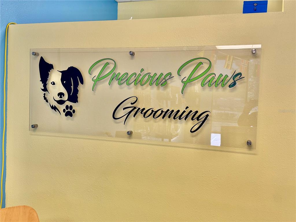 A clear acrylic sign for Precious Paws Grooming is mounted on a light yellow wall — 3283 S JOHN YOUNG PARKWAY, KISSIMMEE, FL