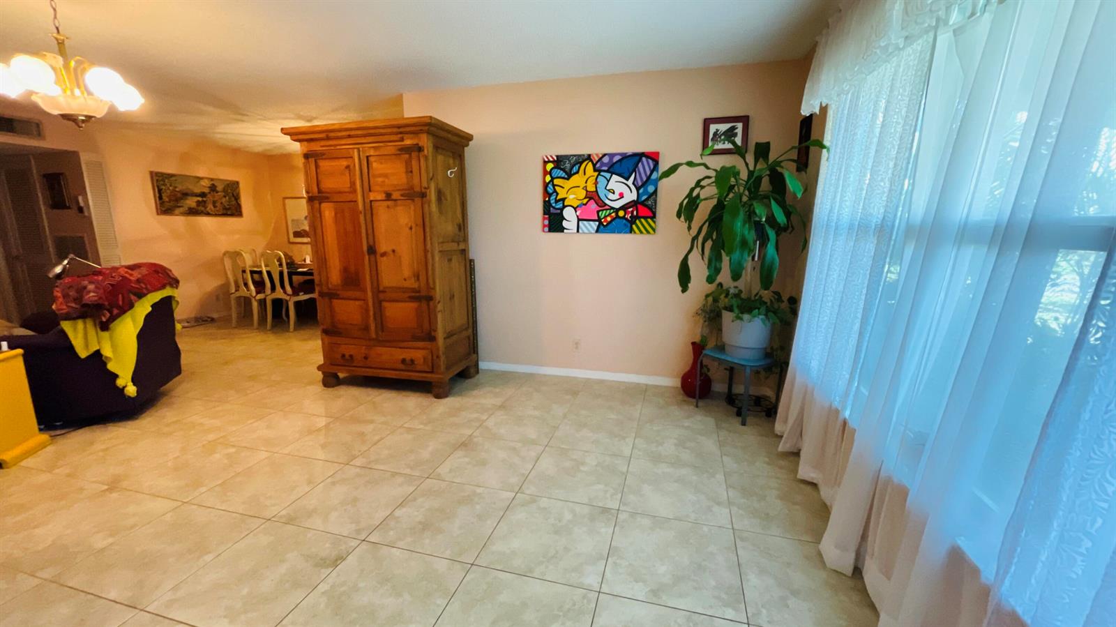 A living room with tile floors, a wooden armoire, colorful wall art, and a large window — 910 South Drive # B, Delray Beach FL 33445, DELRAY BEACH, FL