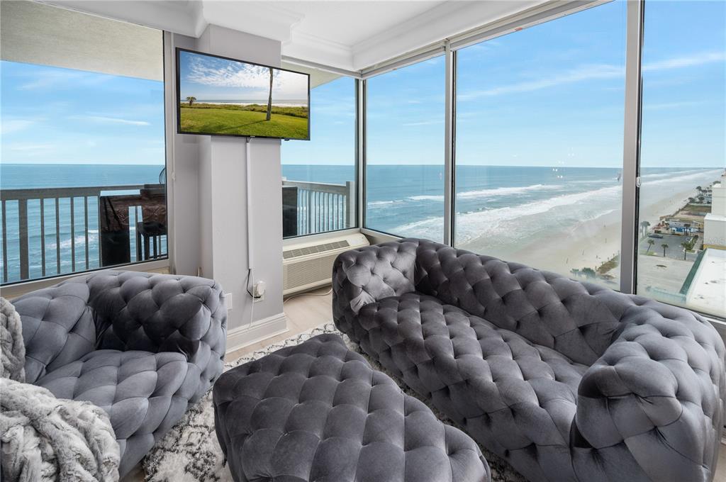 A living room with tufted grey furniture overlooks the ocean through floor-to-ceiling windows — 2700 N ATLANTIC AVENUE UNIT 1210, DAYTONA BEACH, FL