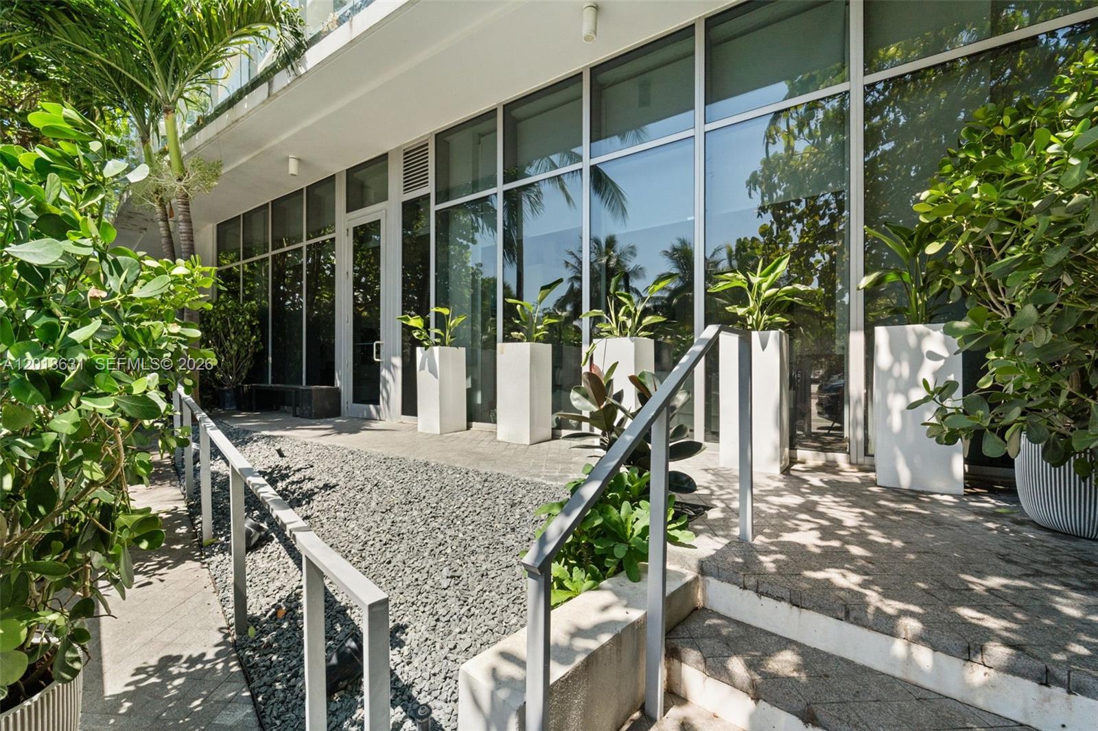 A paved walkway with gravel and potted plants leads to a building with glass walls — 1 Collins Ave # 108, Miami Beach FL 33139, MIAMI BEACH, FL