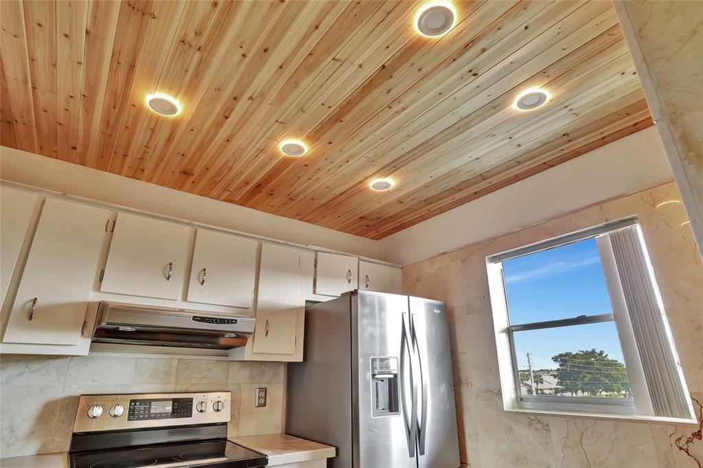 A kitchen features white cabinets, a stainless steel refrigerator, and a wood-planked ceiling with recessed lighting — 4311 Crystal Lake Drive # 421, Pompano Beach FL 33064, POMPANO BEACH, FL
