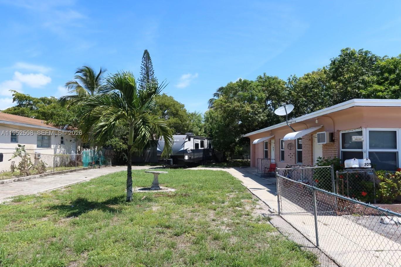 A grassy yard features a palm tree, a parked travel trailer, and a residential building — 205 SE 10th St, Hallandale Beach FL 33009, HALLANDALE BEACH, FL