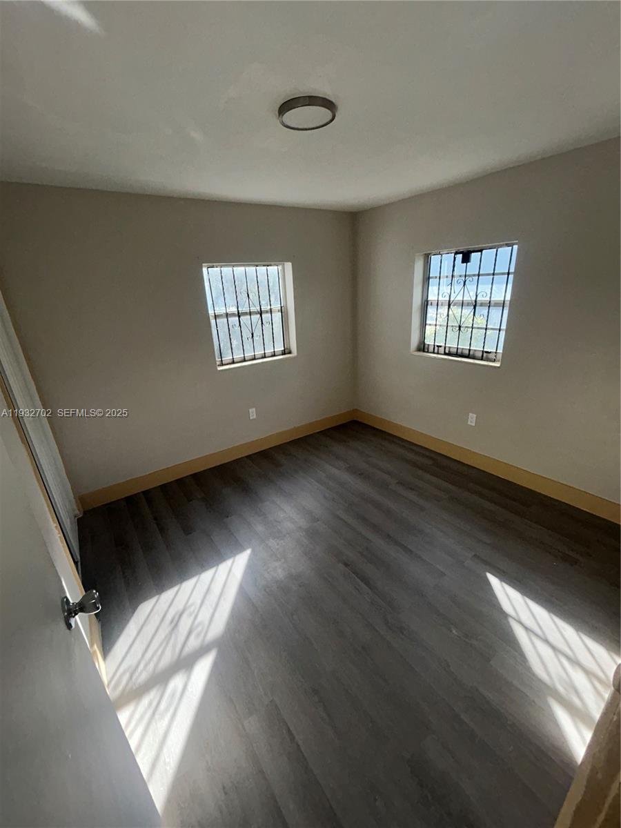 An empty bedroom features gray wood-look flooring, light-colored walls, and two windows with security bars — 1180 NW 64th St # B, Miami FL 33150, MIAMI, FL