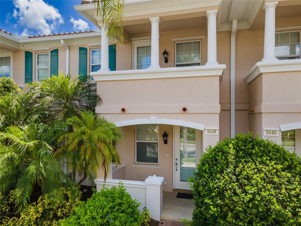 A two-story tan townhouse exterior features a covered entryway, a second-floor balcony, and landscaping — 1648 NAPOLI DRIVE W, SARASOTA, FL