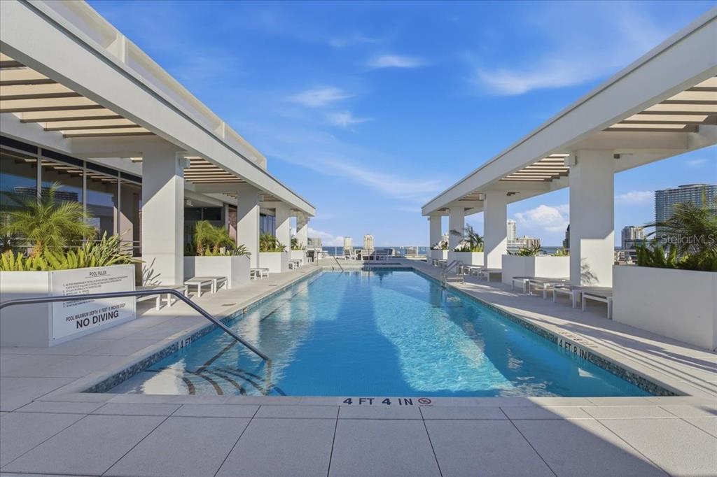 A rectangular outdoor swimming pool is framed by white pergolas on a modern building terrace — 777 3RD AVENUE N UNIT 1202, ST PETERSBURG, FL