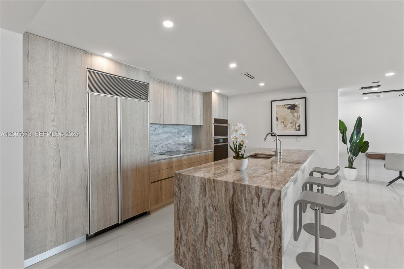 A modern kitchen features light wood cabinetry, a stone waterfall island, and white tile flooring — 1500 Ocean Dr # 1003, Miami Beach FL 33139, MIAMI BEACH, FL