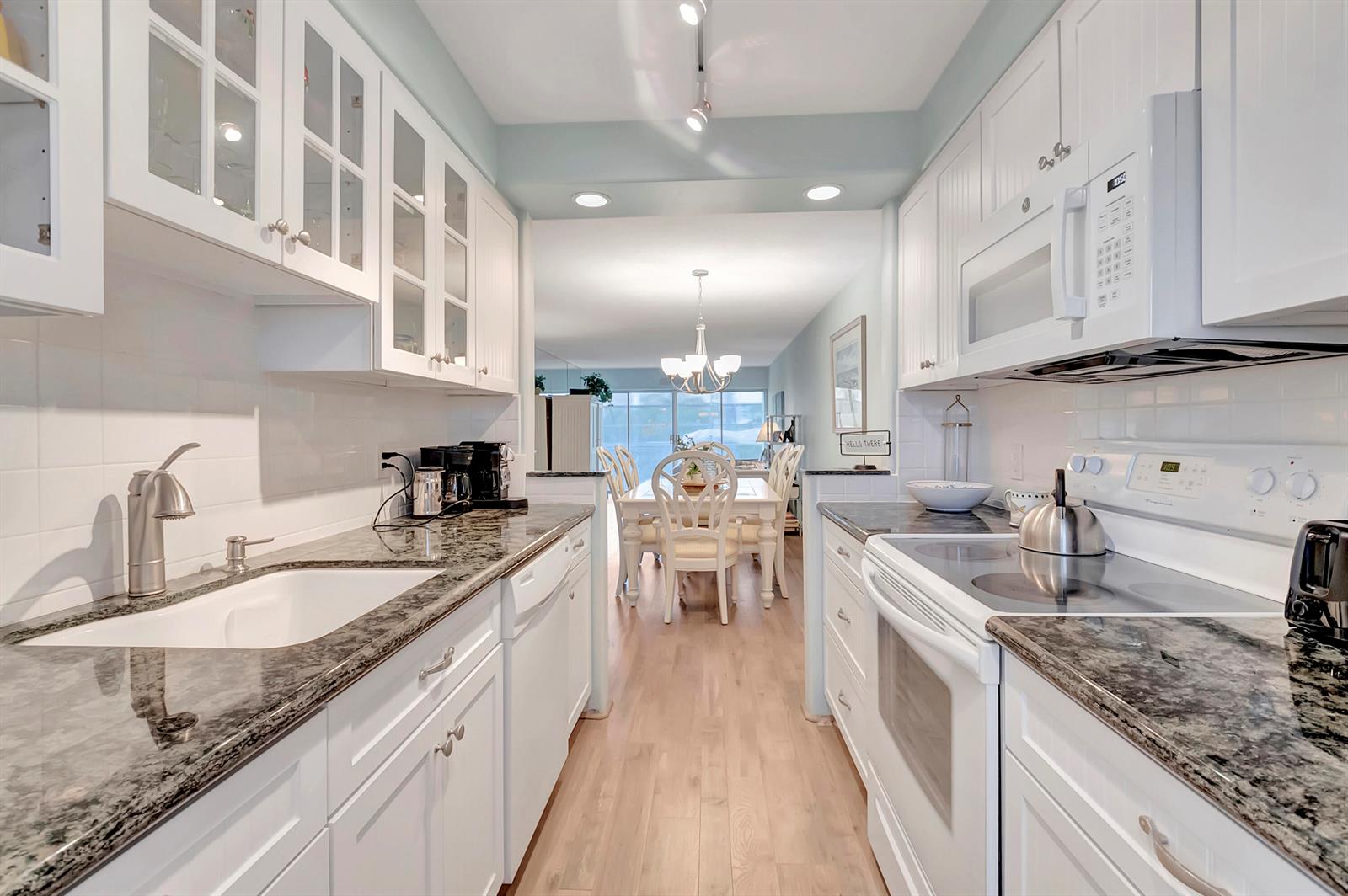 A galley kitchen with white cabinets, granite countertops, and stainless steel appliances leads to a dining area — 1910 S Ocean Boulevard # 416, Delray Beach FL 33483, DELRAY BEACH, FL