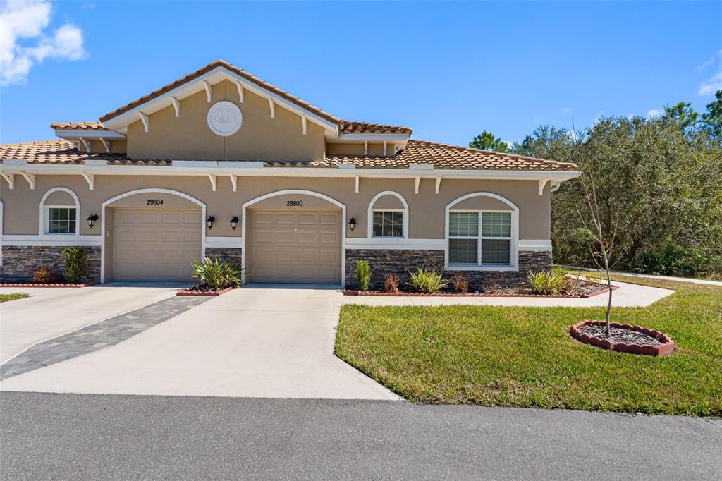 A single-story duplex with tan stucco, stone accents, and two beige garage doors — 29800 ANAND VIHAR DRIVE, WESLEY CHAPEL, FL