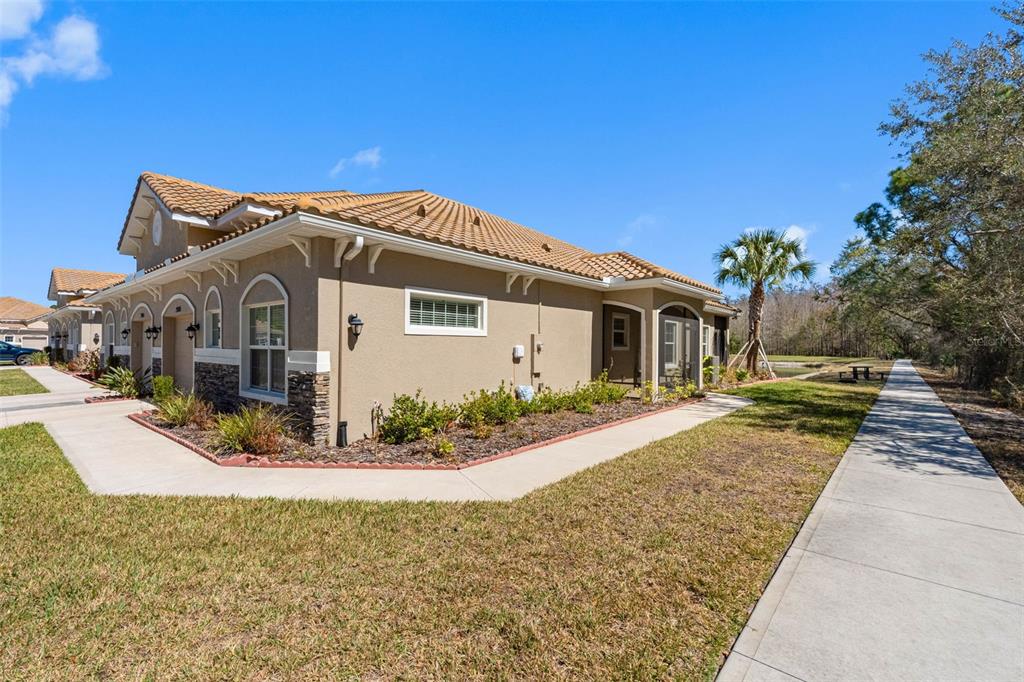 A tan stucco villa with a tiled roof sits beside a sidewalk and a pond — 29800 ANAND VIHAR DRIVE, WESLEY CHAPEL, FL