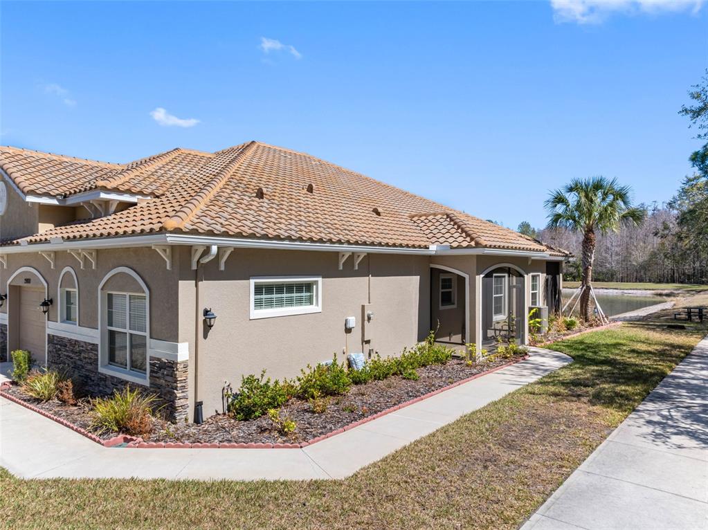 A tan stucco house with a tiled roof sits beside a sidewalk and a pond — 29800 ANAND VIHAR DRIVE, WESLEY CHAPEL, FL