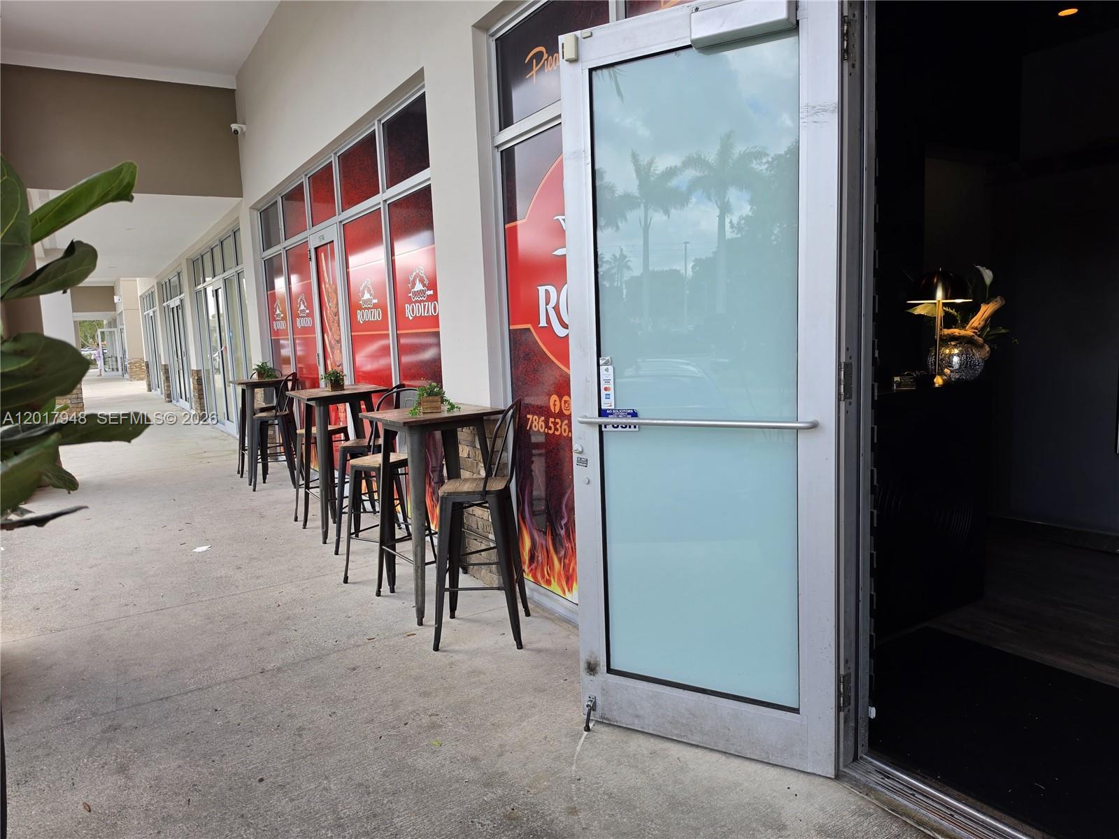 An outdoor seating area with high tables and chairs sits outside a restaurant entrance — 15750 SW 56 ST, Miami FL 33185, MIAMI, FL