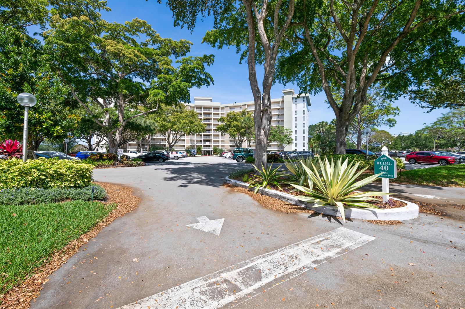 A multi-story apartment building stands behind a parking lot with mature trees and landscaping — 3051 N Course Drive # 211, Pompano Beach FL 33069, POMPANO BEACH, FL