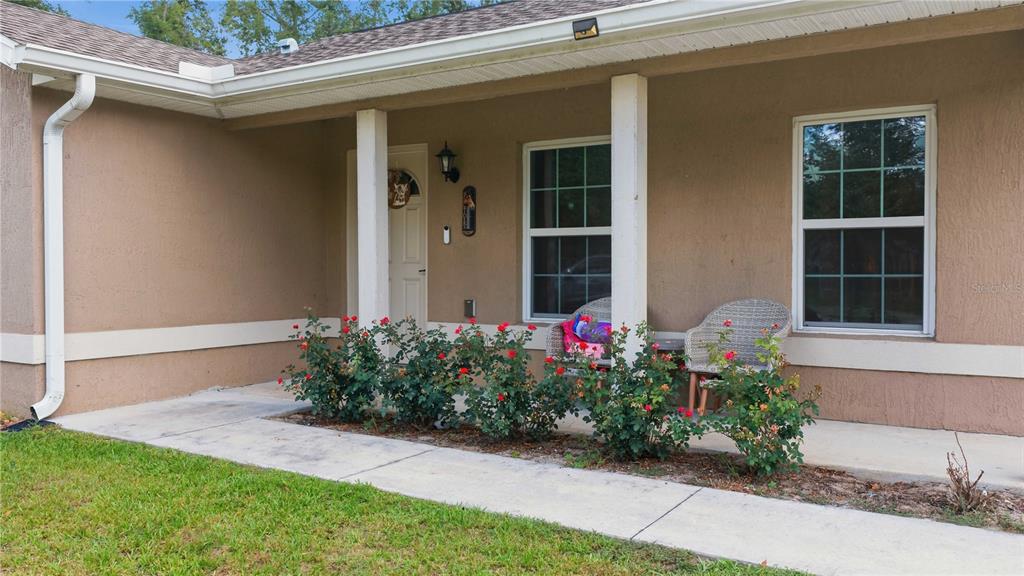 A tan house exterior features a covered porch with two wicker chairs and rose bushes — 14055 SE 43RD TERRACE, SUMMERFIELD, FL