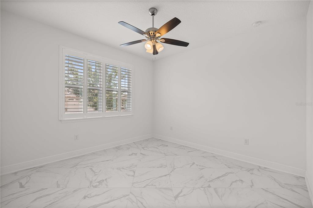 An empty room features white walls, marble-look tile flooring, a ceiling fan, and plantation shutters — 8418 SW 93RD CIRCLE, OCALA, FL