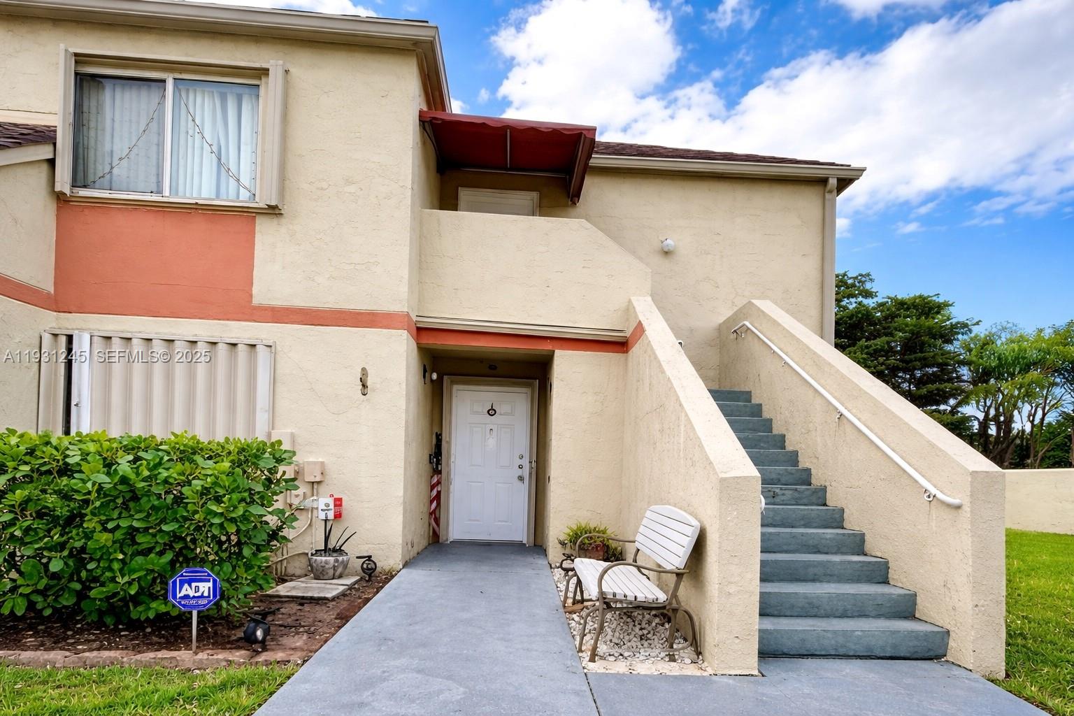 A two-story stucco building features an exterior staircase, a white front door, and a bench — 18810 NW 57th Ave # 102, Hialeah FL 33015, HIALEAH, FL