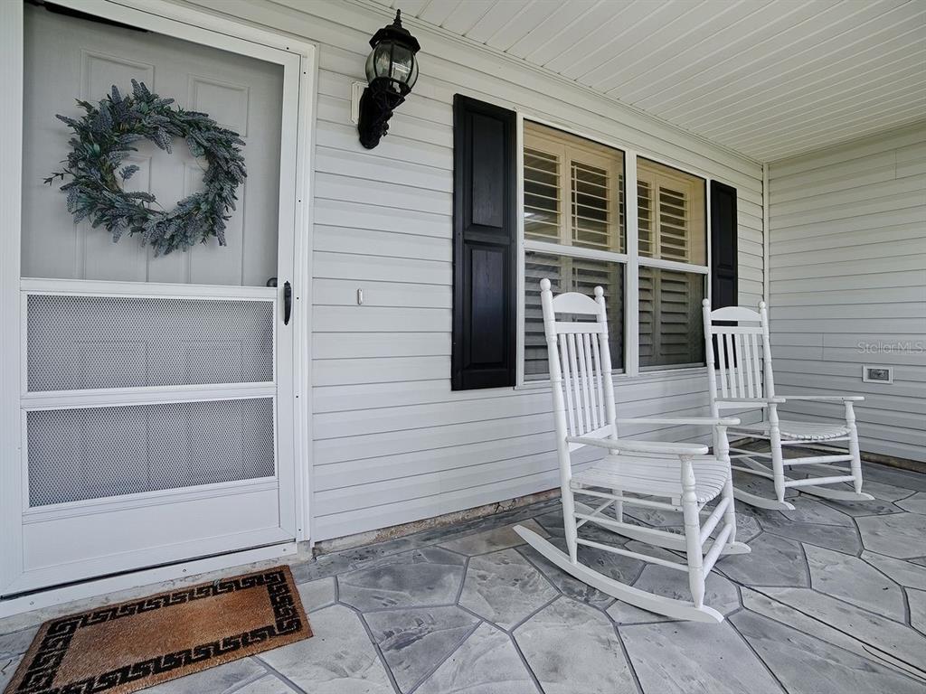 A covered front porch features white siding, two white rocking chairs, and stone-patterned flooring — 2189 CRAWFORD COURT, THE VILLAGES, FL