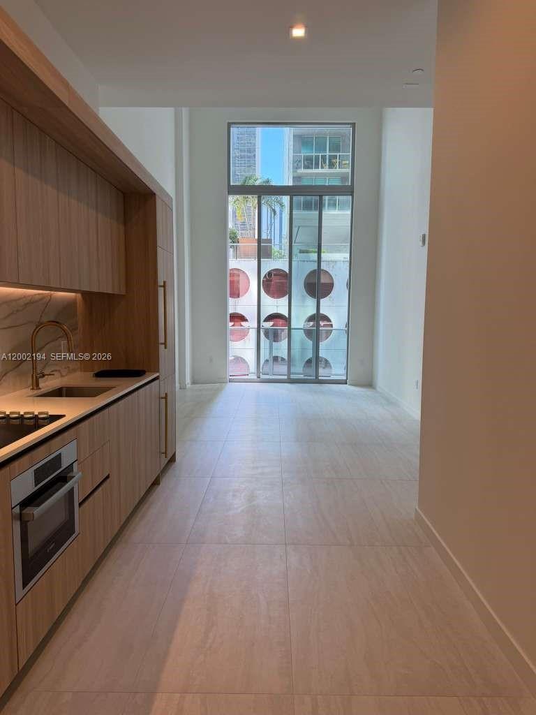 A modern kitchen with light wood cabinetry leads to a floor-to-ceiling windowed balcony — 77 SE 5th street # 1011, Miami FL 33131, MIAMI, FL
