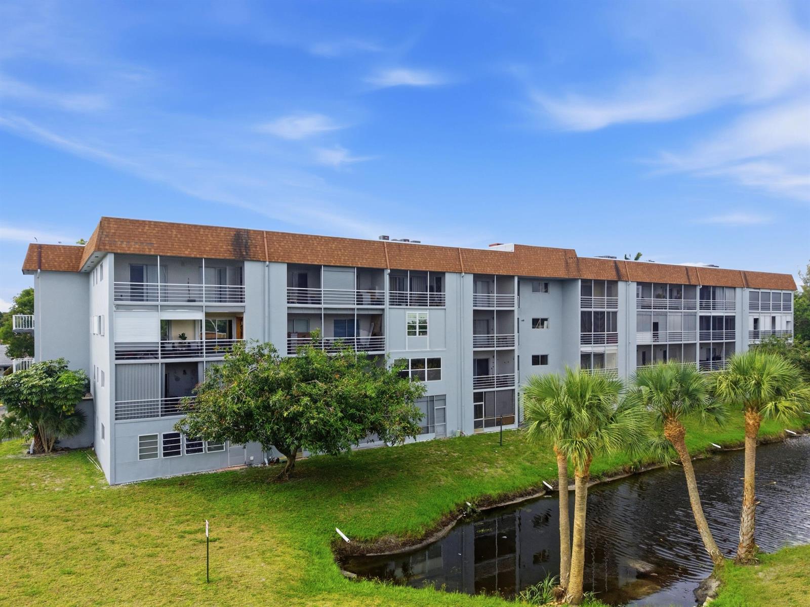 A multi-story apartment building with screened balconies sits beside a small pond and lawn — 6289 Lear Drive # 206, Lake Worth FL 33462, LAKE WORTH, FL