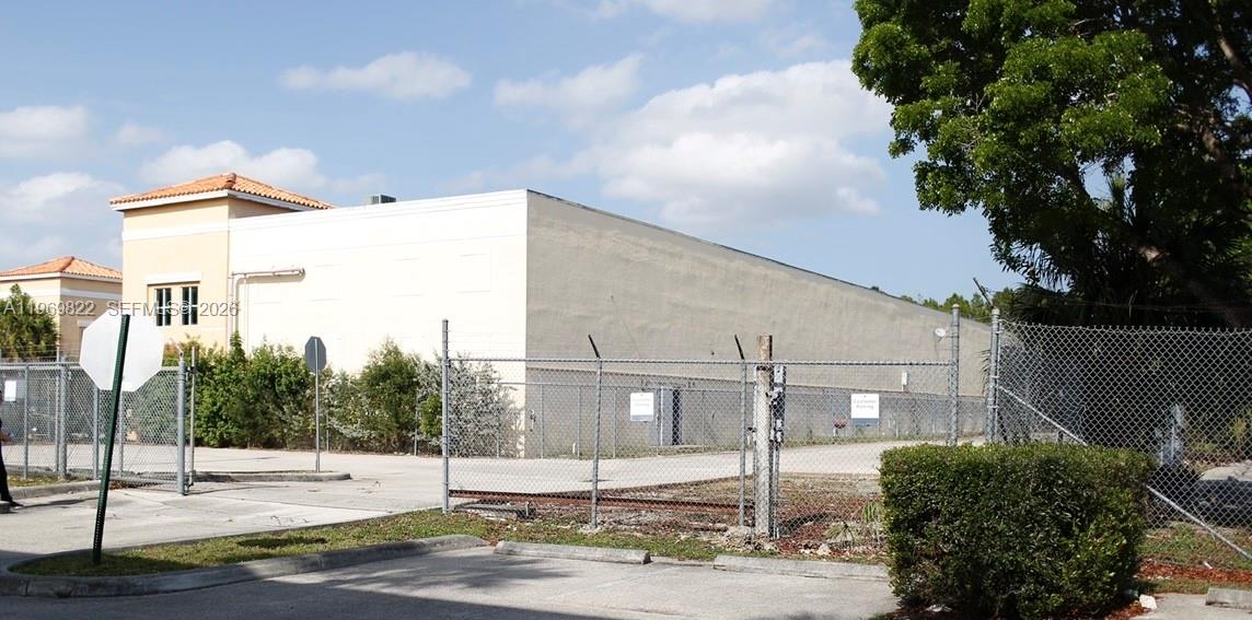 A large, beige, windowless commercial warehouse building is enclosed by a chain-link fence — 12605 SW 134th Ct, Miami FL 33186, MIAMI, FL