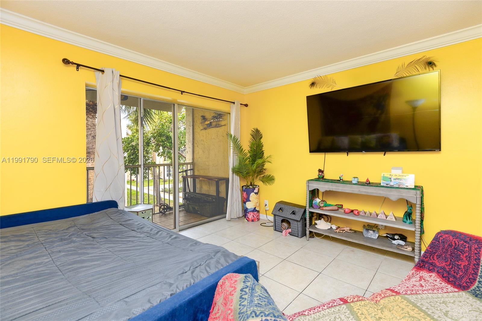 A bedroom features bright yellow walls, a wall-mounted television, a console table, and a balcony — 1428 SE 4th Ave # 240, Deerfield Beach FL 33441, DEERFIELD BEACH, FL