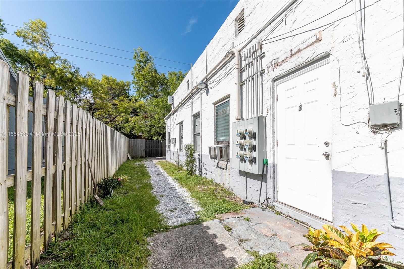 A white building exterior features a gravel path, a wooden fence, and electrical meters — 180 NW 59th St, Miami FL 33127, MIAMI, FL