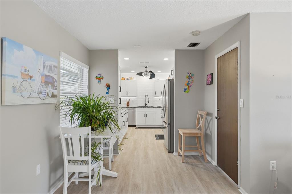 A bright kitchen with white cabinets, light wood-look flooring, and a small dining area — 1127 KING ARTHUR COURT UNIT 316, DUNEDIN, FL
