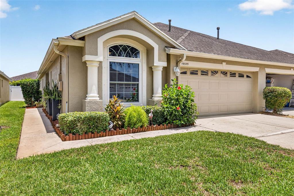 A single-story house exterior features a beige stucco facade, a garage, and landscaped front yard — 19355 HASKELL PLACE, LAND O LAKES, FL