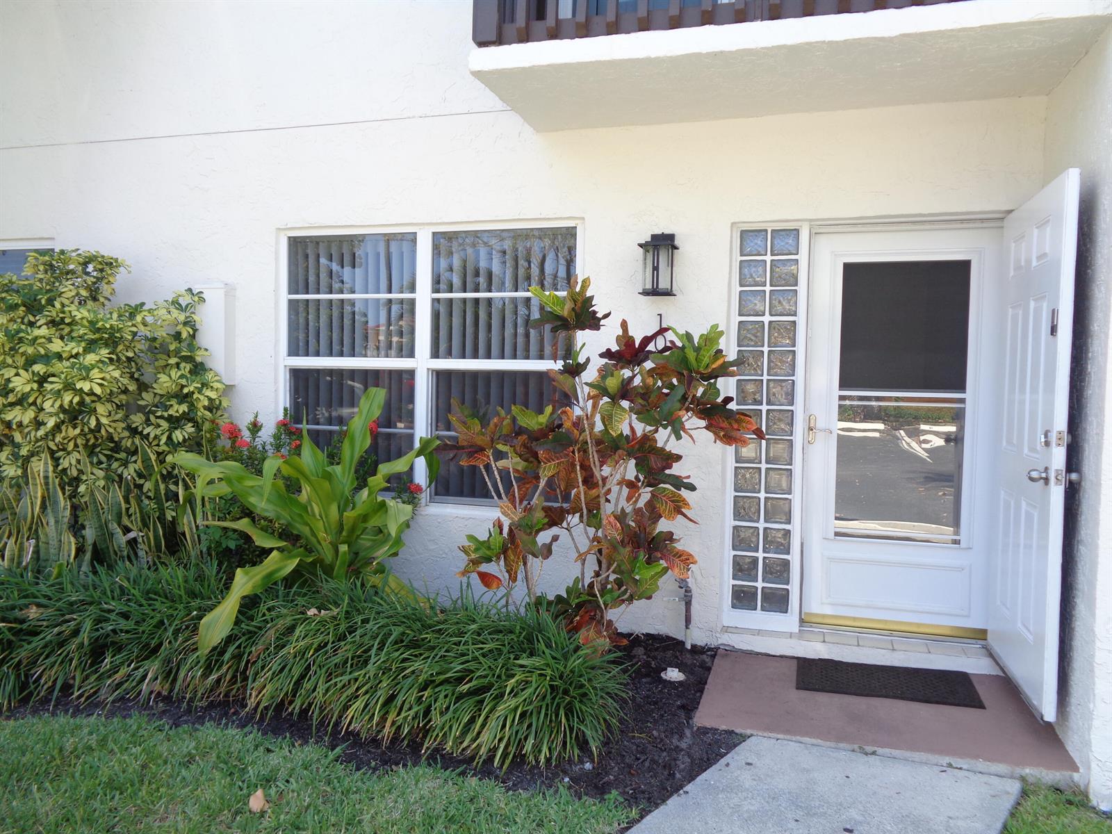 Exterior view of a white building entrance with a glass-paneled door and landscaped garden beds — 1917 NE 3rd Street # 103, Deerfield Beach FL 33441, DEERFIELD BEACH, FL