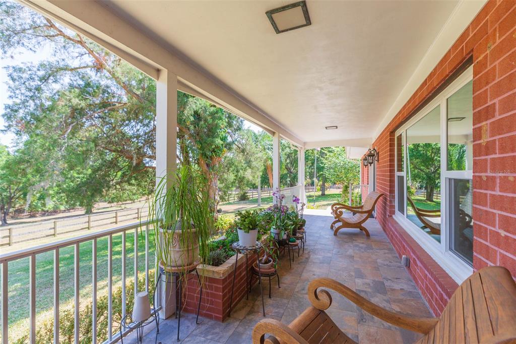A covered brick porch features tiled flooring, potted plants, and wooden chairs overlooking a grassy yard — 34211 BLANTON ROAD, DADE CITY, FL
