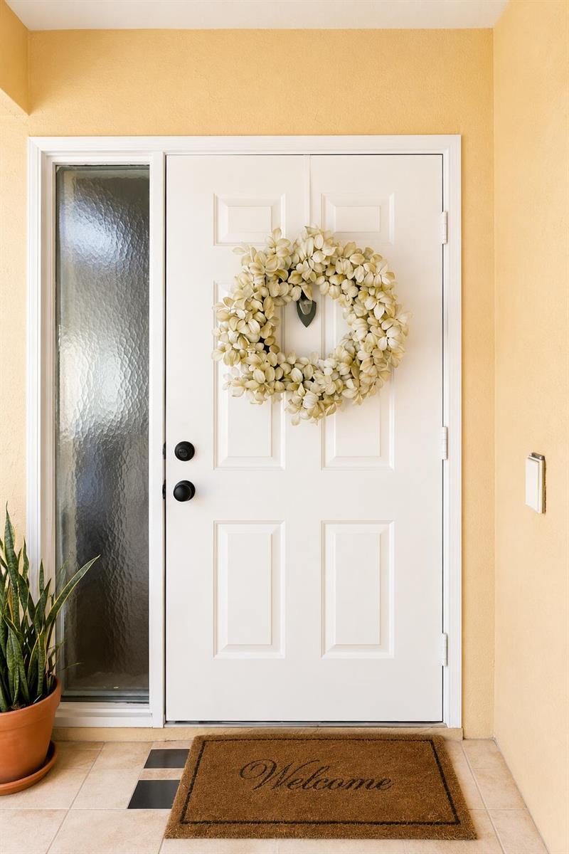 A white front door with a floral wreath and a welcome mat sits on a porch — 179 Waterside Drive # 179, Hypoluxo FL 33462, HYPOLUXO, FL