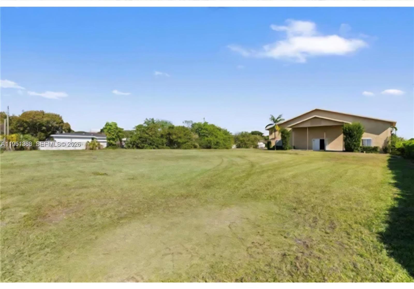 A large, grassy vacant lot sits in front of a tan building under a blue sky — 21571 SW 110th Ave, Miami FL 33189, MIAMI, FL